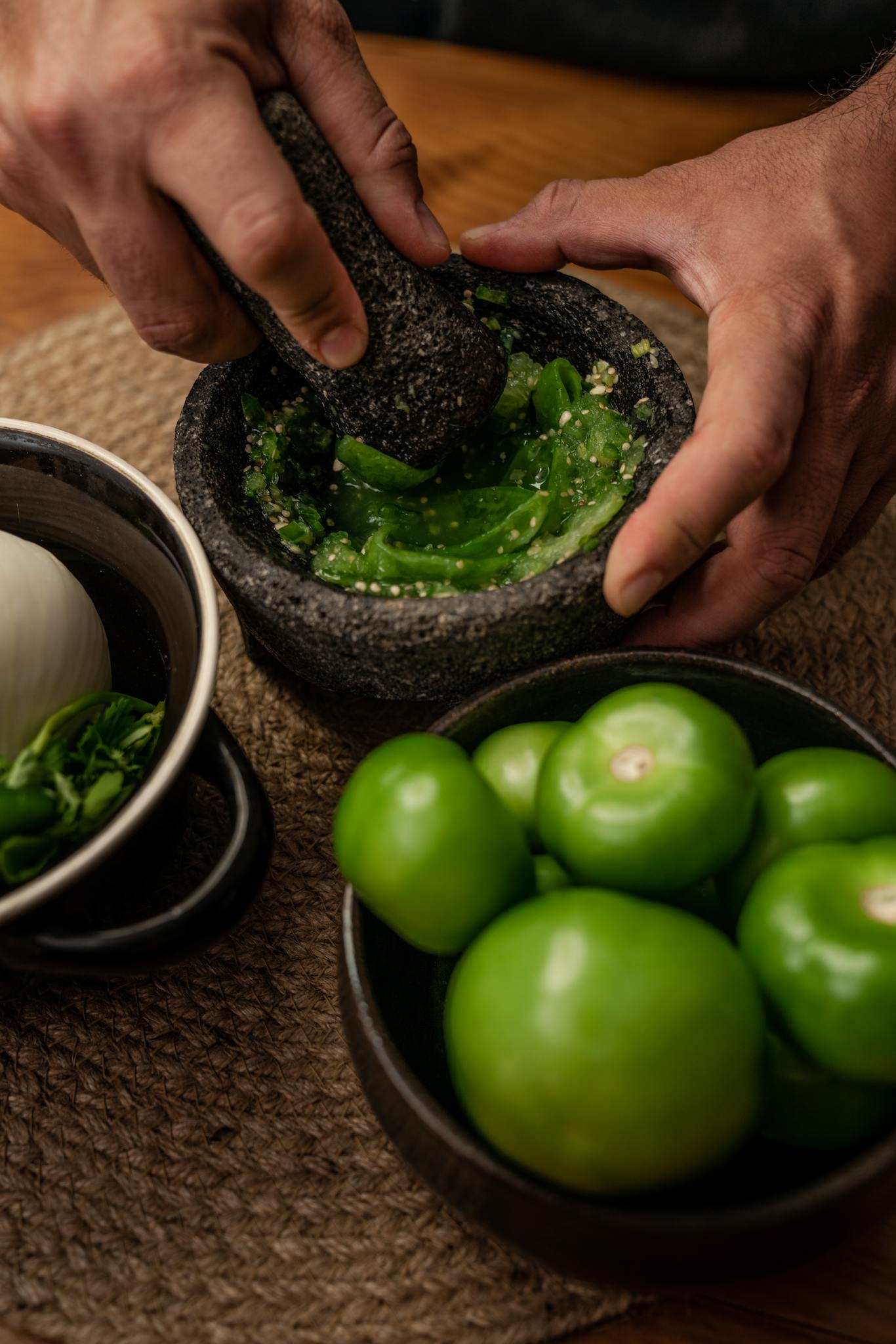 Hands using mortar and pestle to prepare green salsa with fresh ingredients including tomatillos and jalapeños.